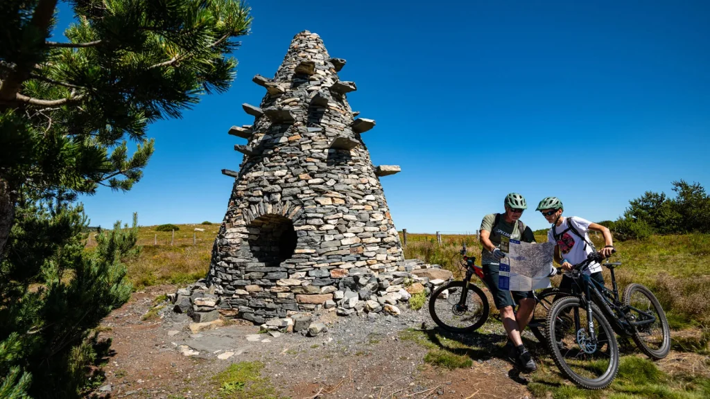 The Ardeche Great Mountain Bike Route, Two men on mountain bikes observing a map in front of the water tower, a work of the "Partage des eaux" by Gilles Clément, in summer