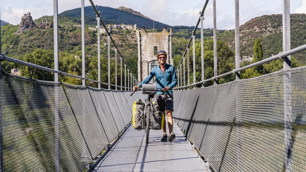 Homme en itinérance, passant, avec son vélo sur la passerelle Himalayenne du village de caractère de Rochemaure, en hiver, pour rejoindre la ViaRhôna